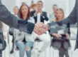Close-up of two businessmen shaking hands in a Custom Development meeting in Salt Lake City, UT, with a blurred group applauding in the background.