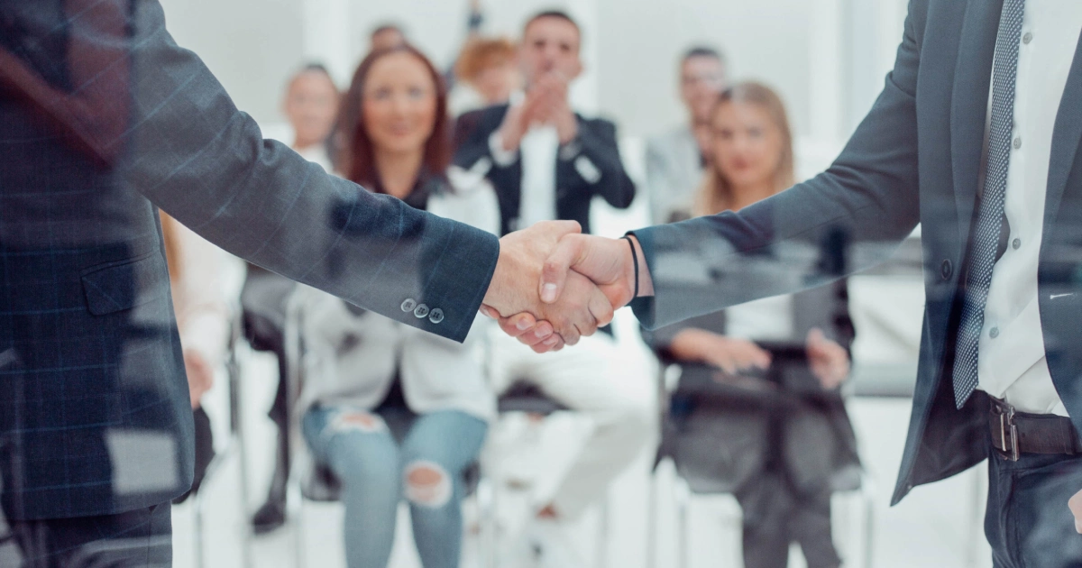 Close-up of two businessmen shaking hands in a Custom Development meeting in Salt Lake City, UT, with a blurred group applauding in the background.