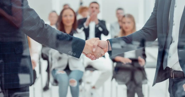 Close-up of two businessmen shaking hands in a Custom Development meeting in Salt Lake City, UT, with a blurred group applauding in the background.